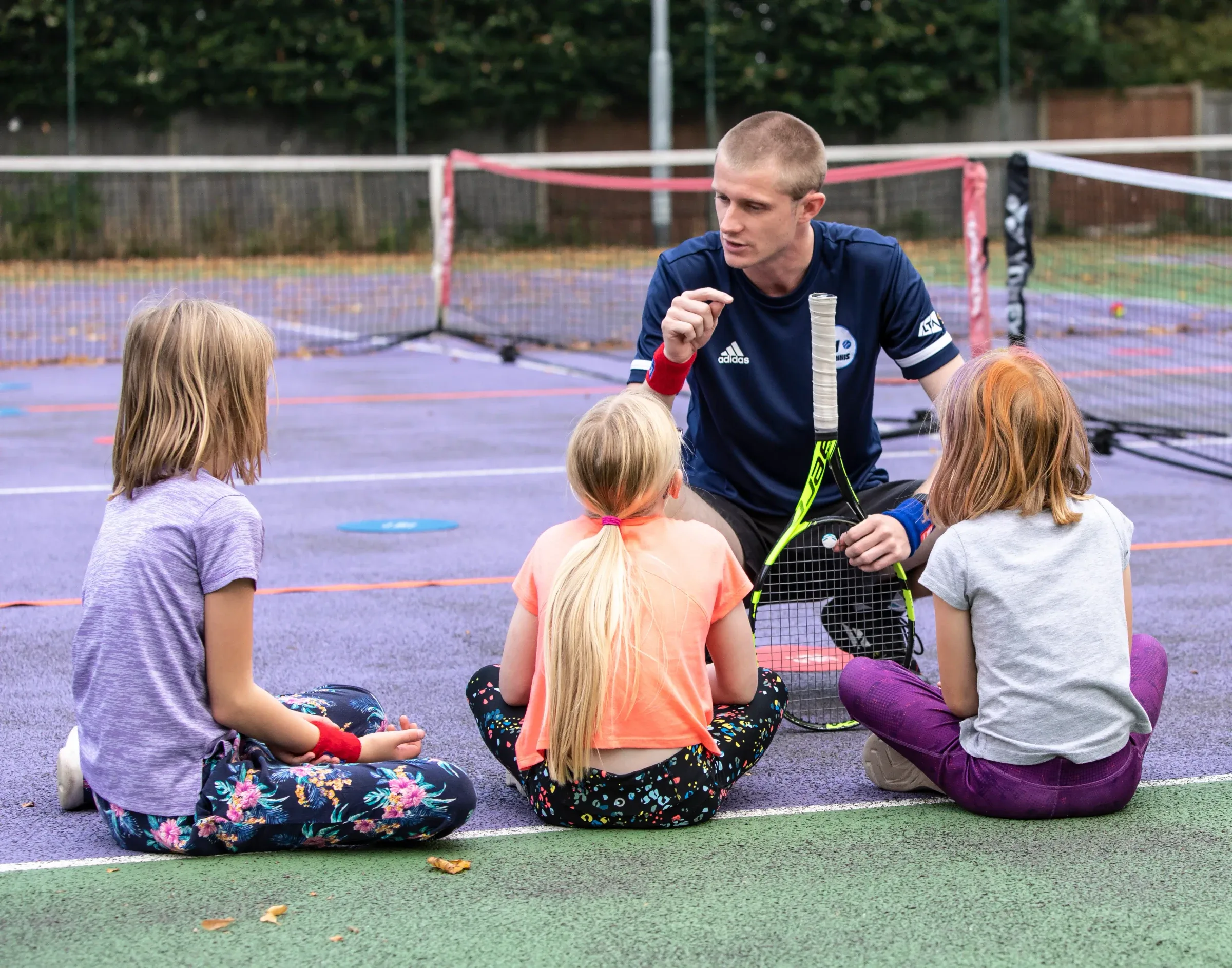 An MH Tennis coach kneeling on court, talking with three young girls sitting cross-legged