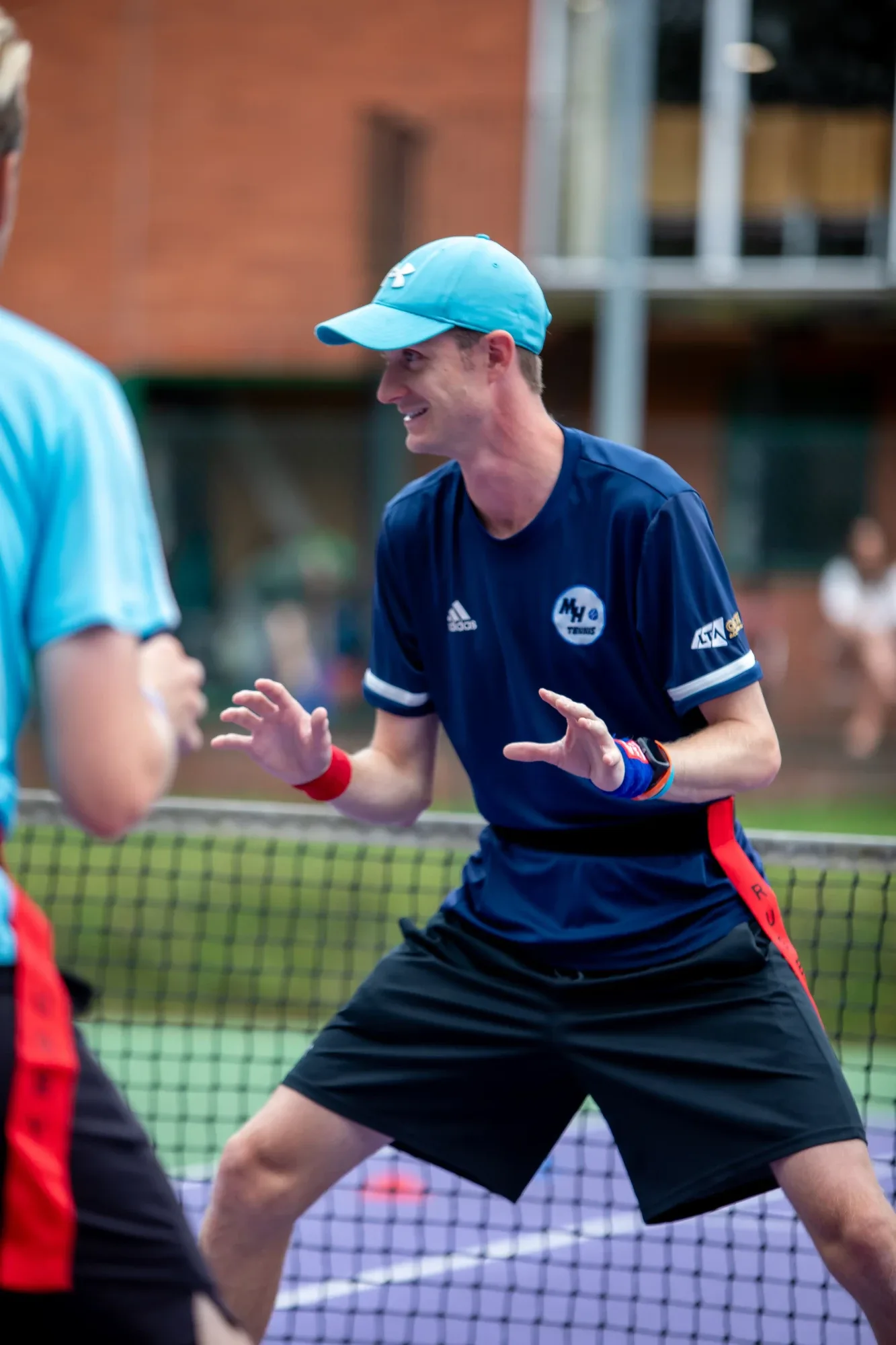 An MH Tennis coach in a cap, smiling and ready at the net during a session